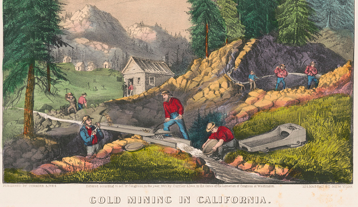 Gold miners shoveling sand from stream into sluice while one miner pans for gold in the same stream, small building and mountains in the background.